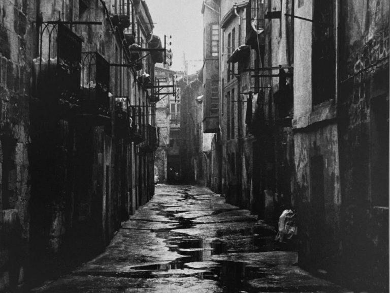 Calle estrecha y empedrada, con edificios antiguos y luces apagadas, tras la lluvia.